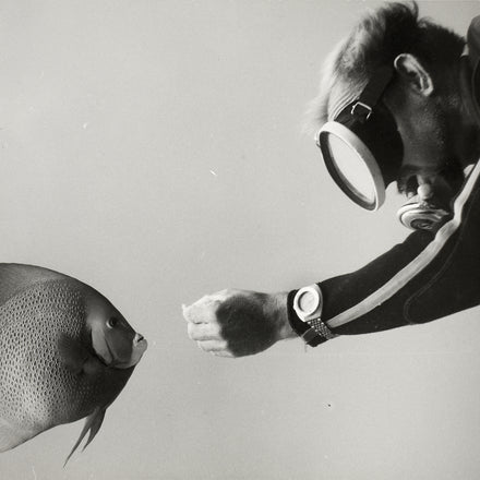 A black and white photo of a diver reaching out toward a fish, the reaching hand's wrist wears a DOXA watch