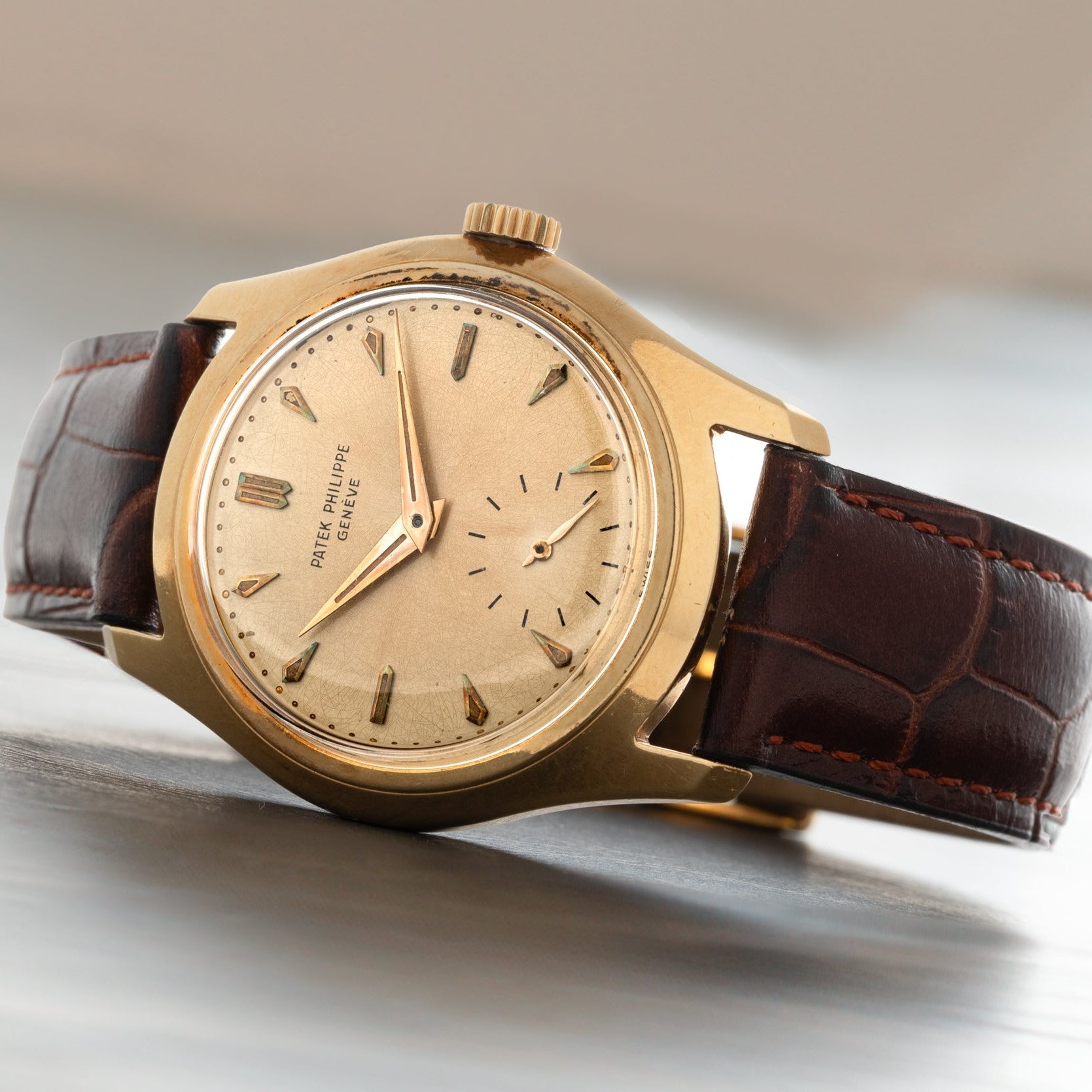 The Patek Philippe Calatrava features a gold case, brown leather strap, cream dial, and gold hour markers, elegantly displayed on a light surface.