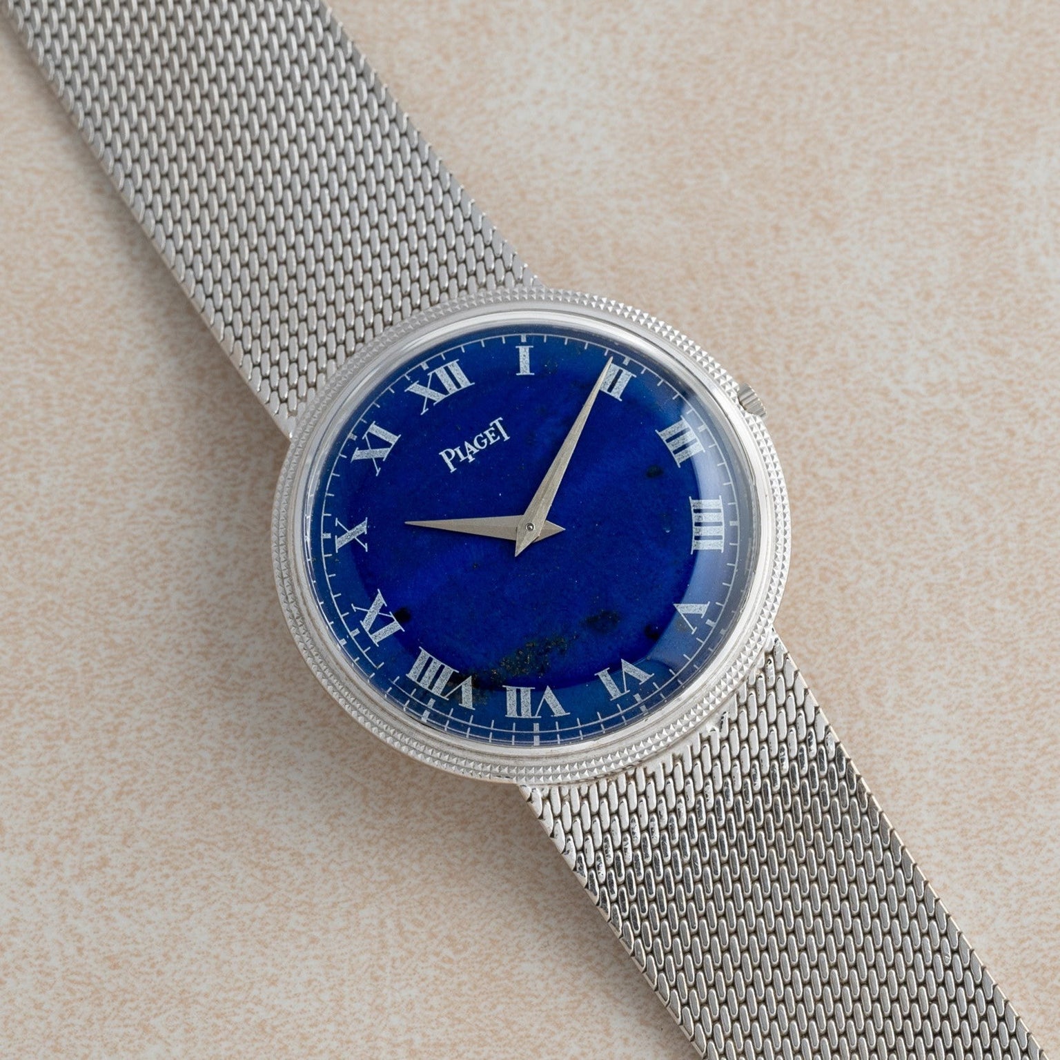 The Piaget Lapis Lazuli Dress Watch features a silver mesh band and blue face with Roman numerals, displayed on a light beige surface.