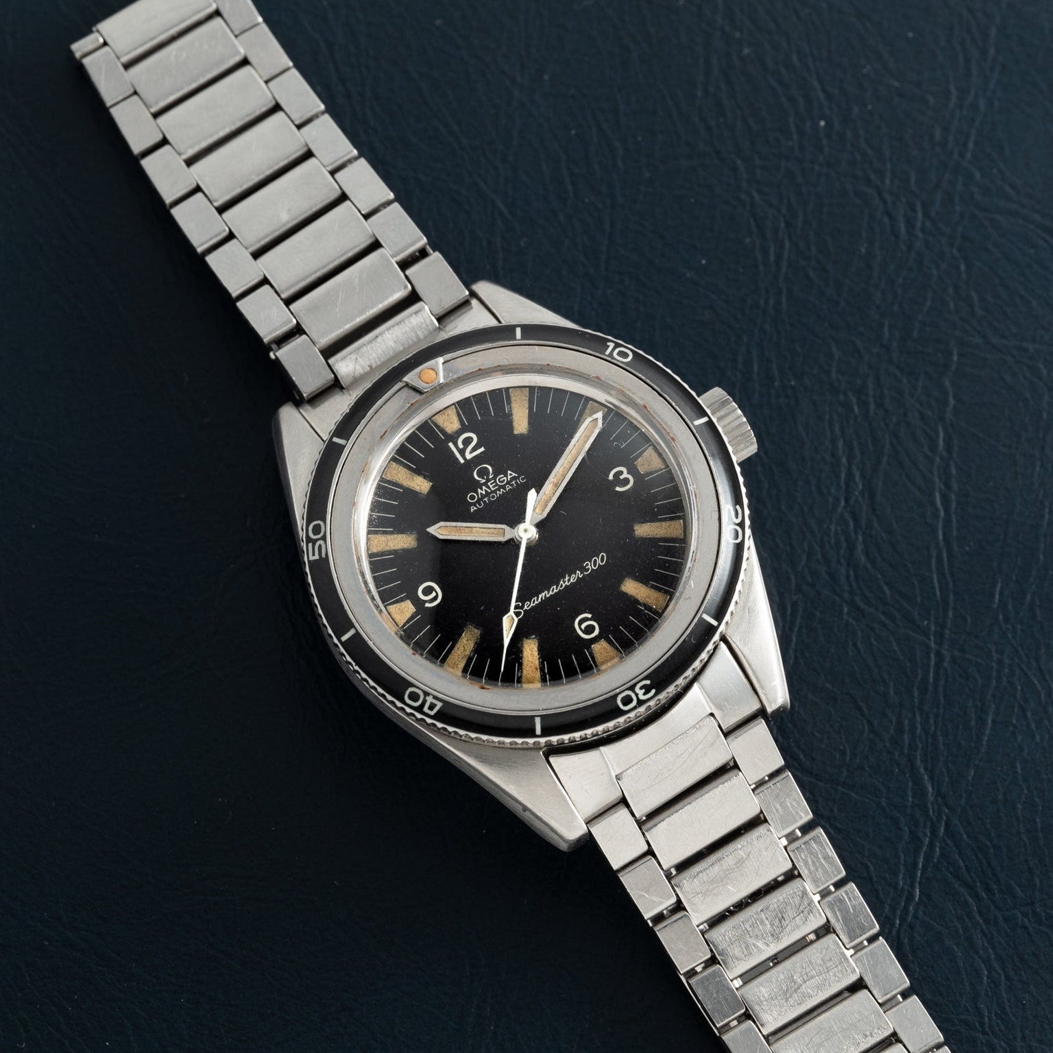 An OMEGA Seamaster 300 watch with a black dial and stainless steel band displayed on a dark textured surface.