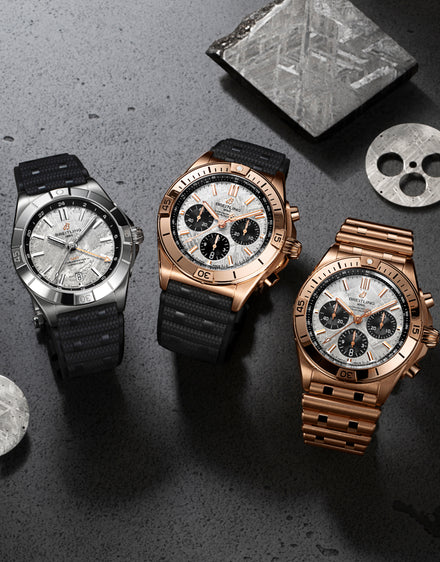 Three luxury chronograph watches with black and gold bands displayed on a stone surface with metal pieces.
