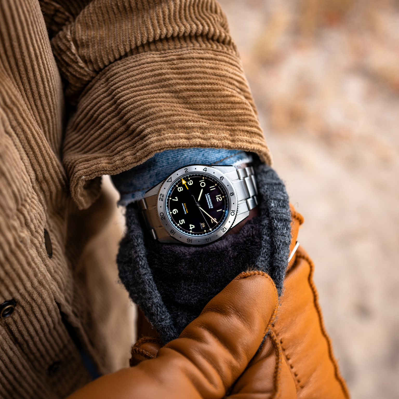 A person in a brown corduroy jacket, blue shirt, and tan leather gloves showcases the Longines Spirit Zulu Time Limited Edition For Hodinkee wristwatch by Longines, featuring a black dial with yellow accents.