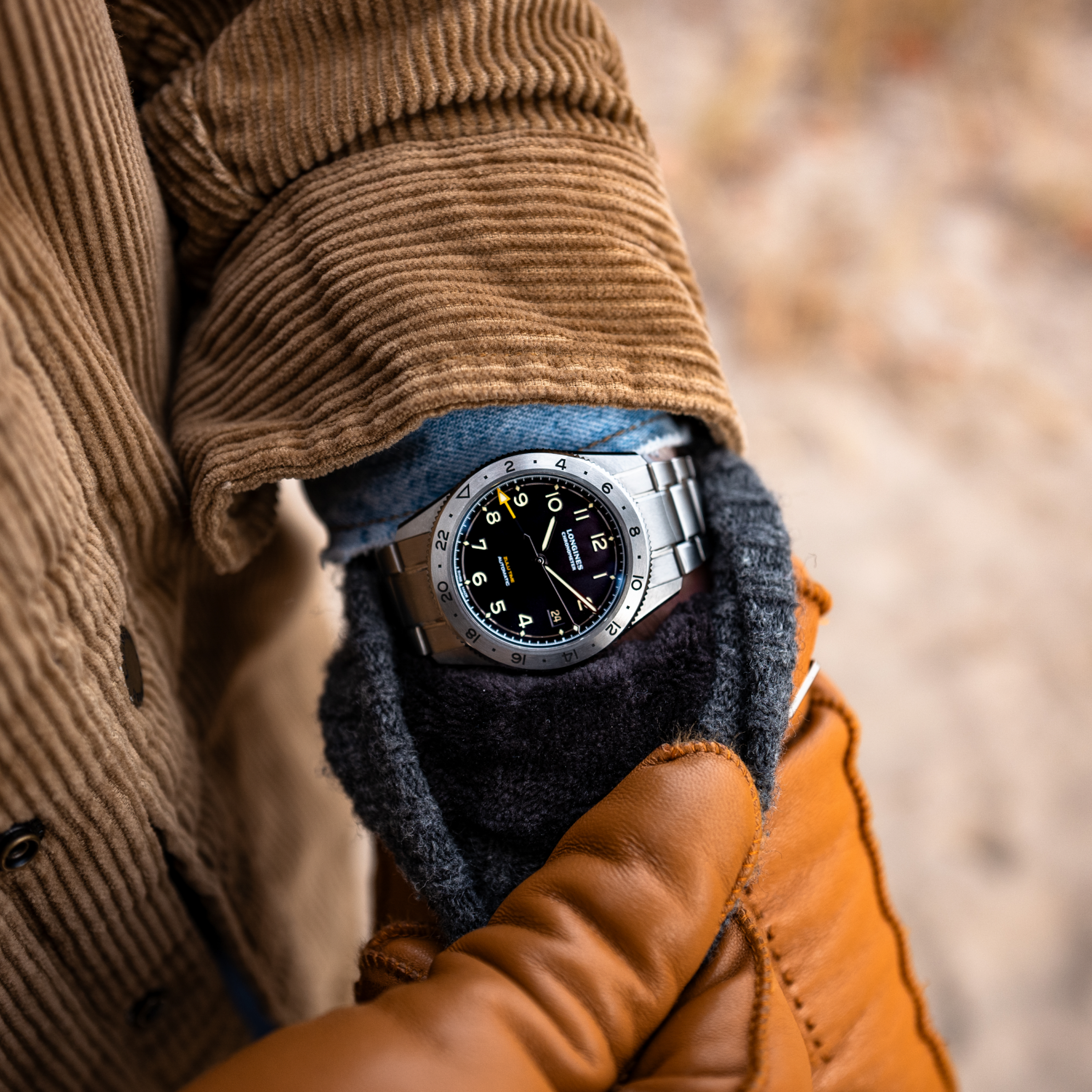 A person in a brown corduroy jacket, blue shirt, and tan leather gloves showcases the Longines Spirit Zulu Time Limited Edition For Hodinkee wristwatch by Longines, featuring a black dial with yellow accents.