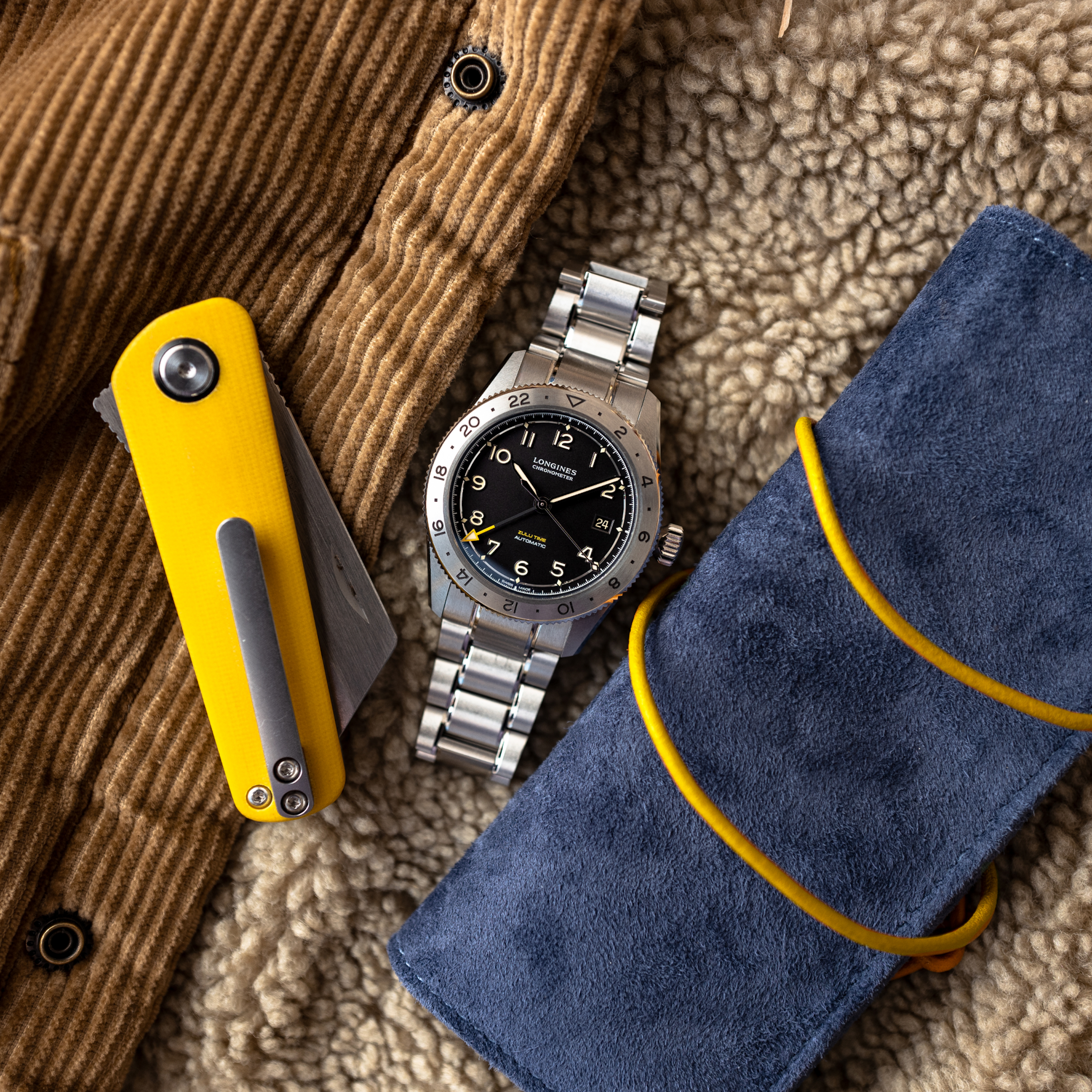 A Longines Spirit Zulu Time Limited Edition For Hodinkee wristwatch, a yellow pocketknife, and a rolled-up blue suede case rest on a tan corduroy and shearling jacket.
