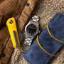 A Longines Spirit Zulu Time Limited Edition For Hodinkee wristwatch, a yellow pocketknife, and a rolled-up blue suede case rest on a tan corduroy and shearling jacket.