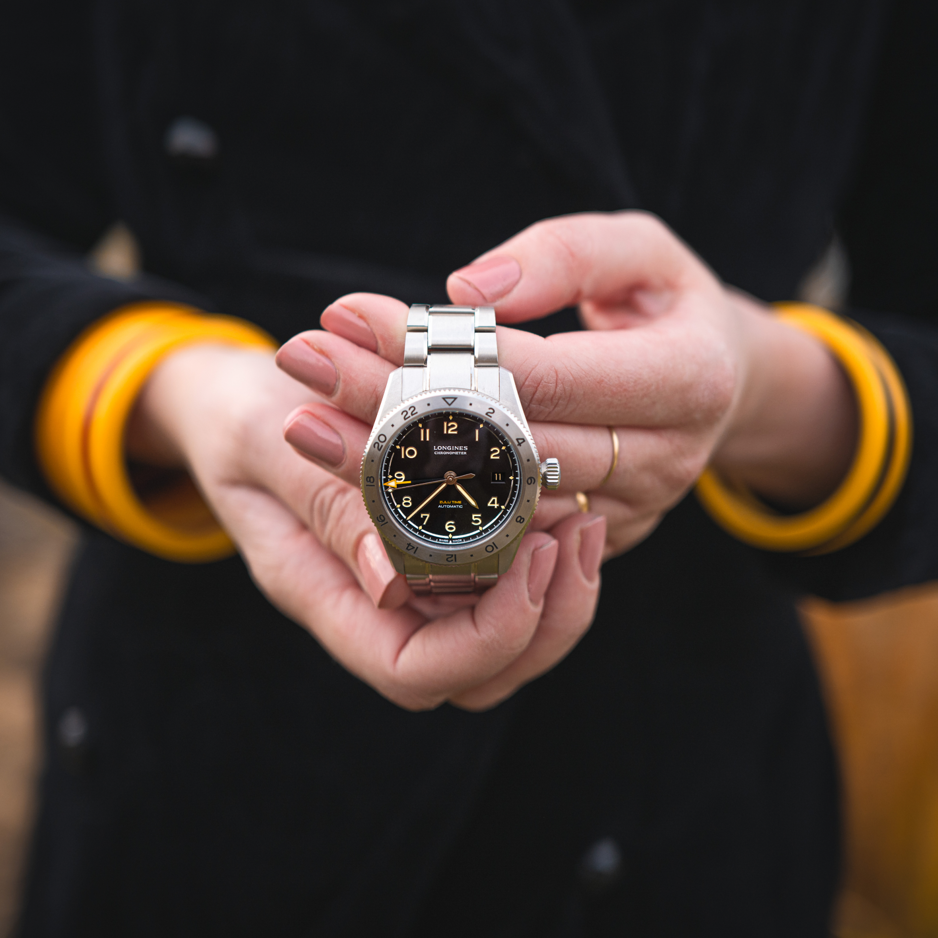 A person in a black coat with yellow cuffs holds the Longines Spirit Zulu Time Limited Edition For Hodinkee wristwatch in both hands, featuring Grade 5 Titanium, a black dial, and classic numbering.