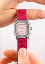 Pink watch with diamond-studded face held between fingers against a light background