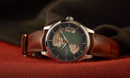 A Hamilton Jazzmaster watch with a brown leather strap and gray-green dial with cutouts on a red fabric surface with a folded brown fabric background