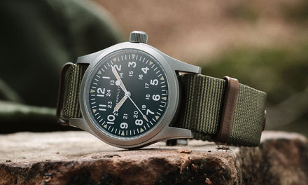 A Hamilton watch with a green khaki fabric strap and a  silver tone bezel, sitting outdoors on a rock surface