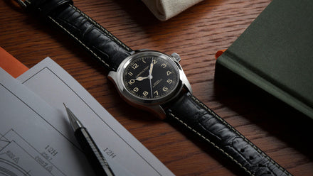 A wristwatch with a black leather strap rests on a wooden desk next to papers, a pen, and a green book.