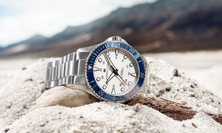 A Hamilton watch with a silver-tone strap and white dial with a blue numbered bezel sitting in sand with a rugged mountainous background