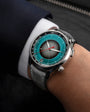 Silver chronograph watch with teal dial, central circular window, red hands, and dark leather strap worn on wrist.