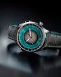 Silver chronograph watch with teal dial, black leather strap, and dual time zone subdial.