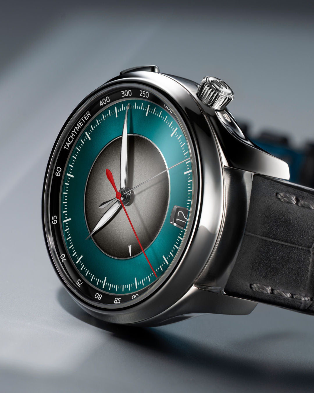 Silver chronograph watch with teal dial, center subdial, red chronograph hand, and black leather strap.