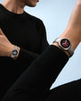 Man wearing black shirt and pants with two watches on wrists, one with dark face and one with burgundy face against blue sky.