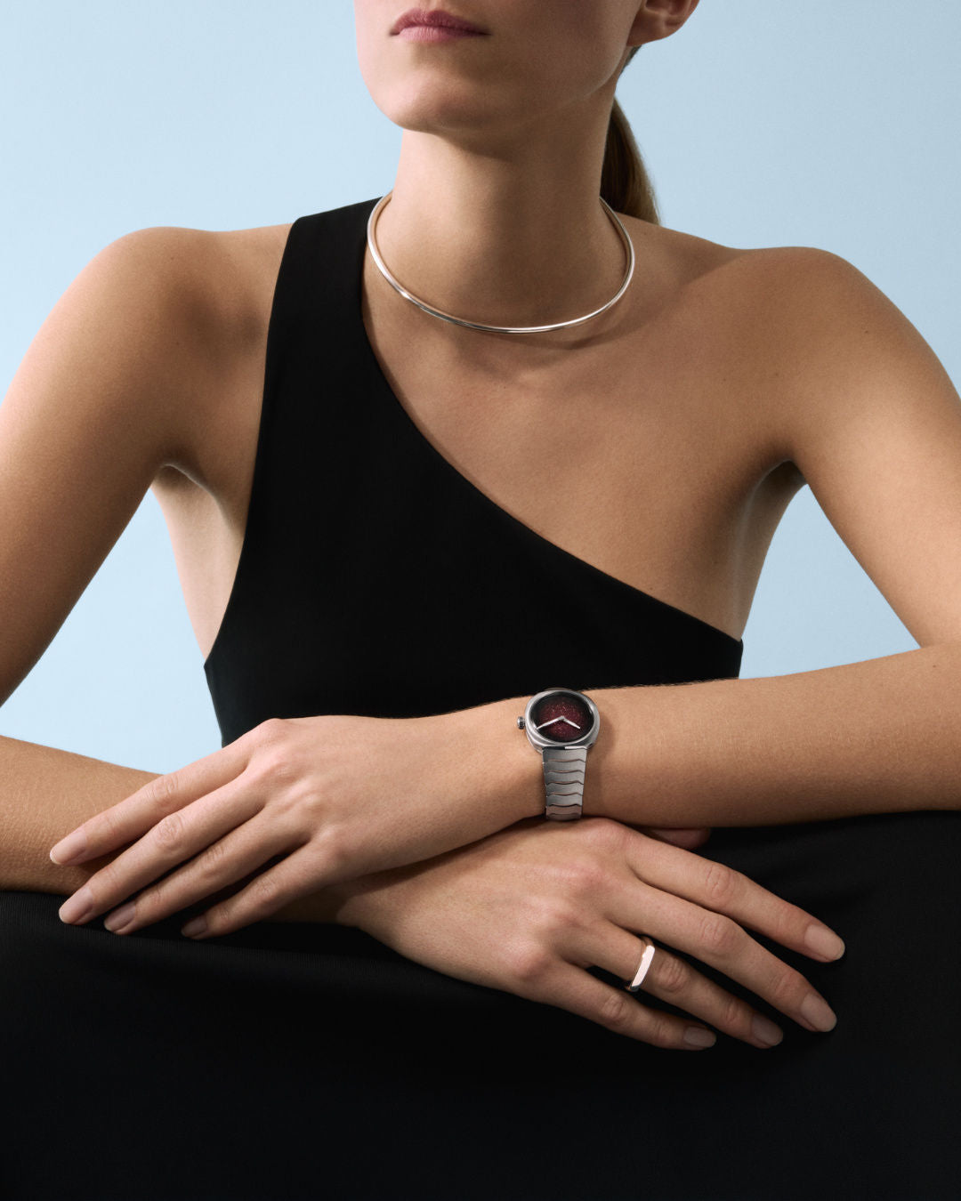 Woman wearing black one-shoulder top with silver watch on wrist and silver neck collar against blue background.