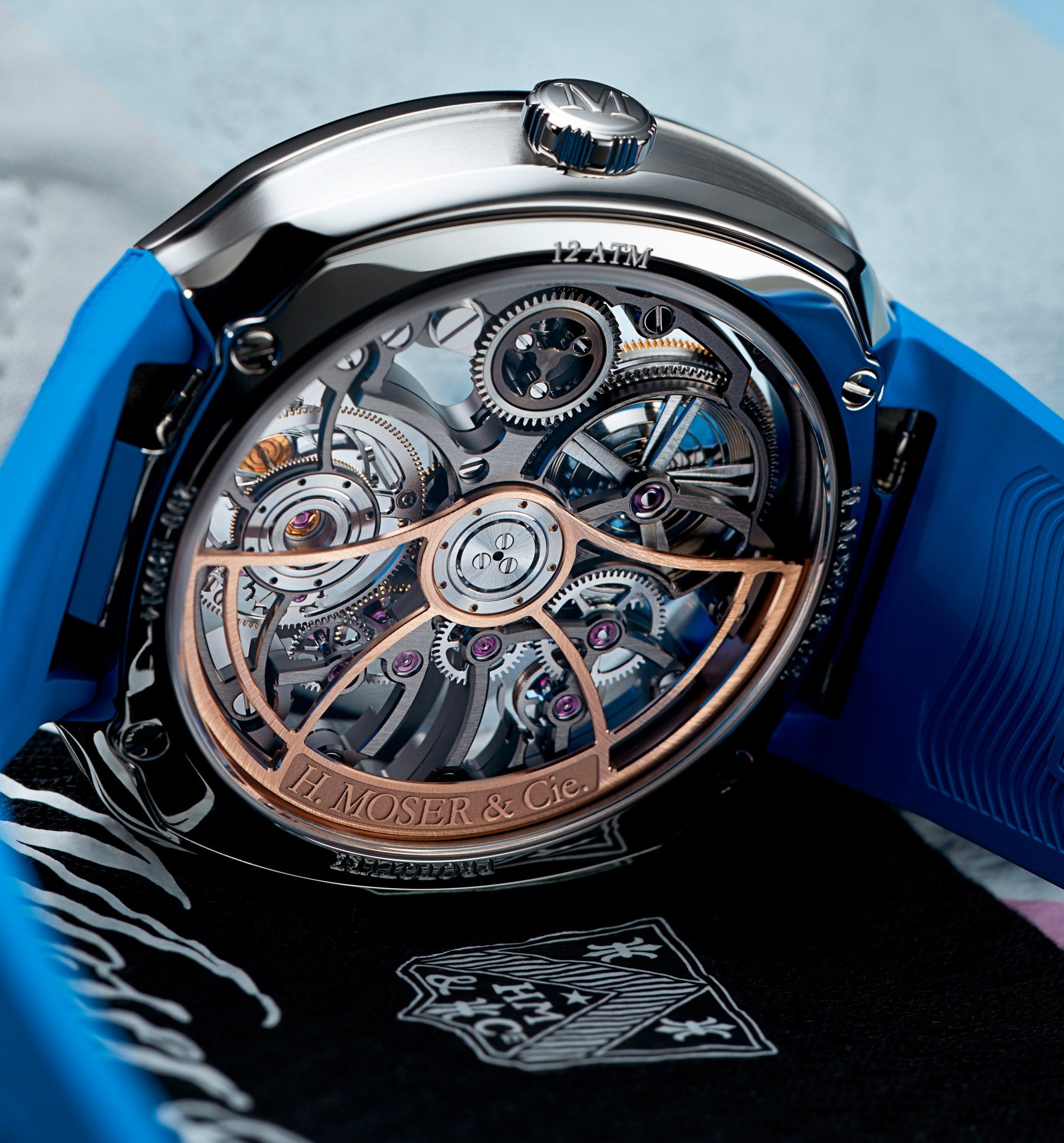 Luxury watch with exposed rose gold and silver mechanical movement, polished steel case, blue rubber strap, and visible H. Mo