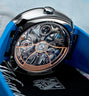 Luxury watch with exposed rose gold and silver mechanical movement, polished steel case, blue rubber strap, and visible H. Mo