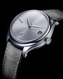 Tantalum watch with brushed silver dial, three hands, date window, and gray leather strap against black background.