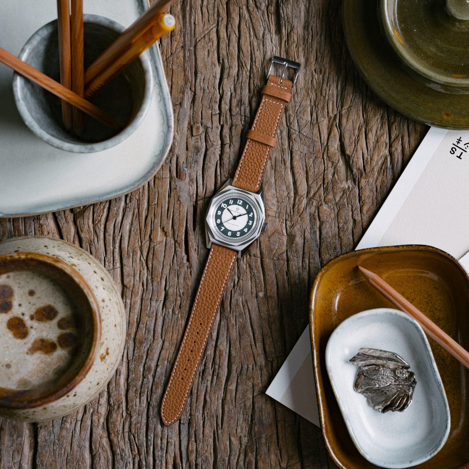 The Merci INSTRUMENTS Beaumarchais H02 Limited Edition for Hodinkee by Merci rests on a rustic wooden table, surrounded by pottery, chopsticks, and scattered papers.