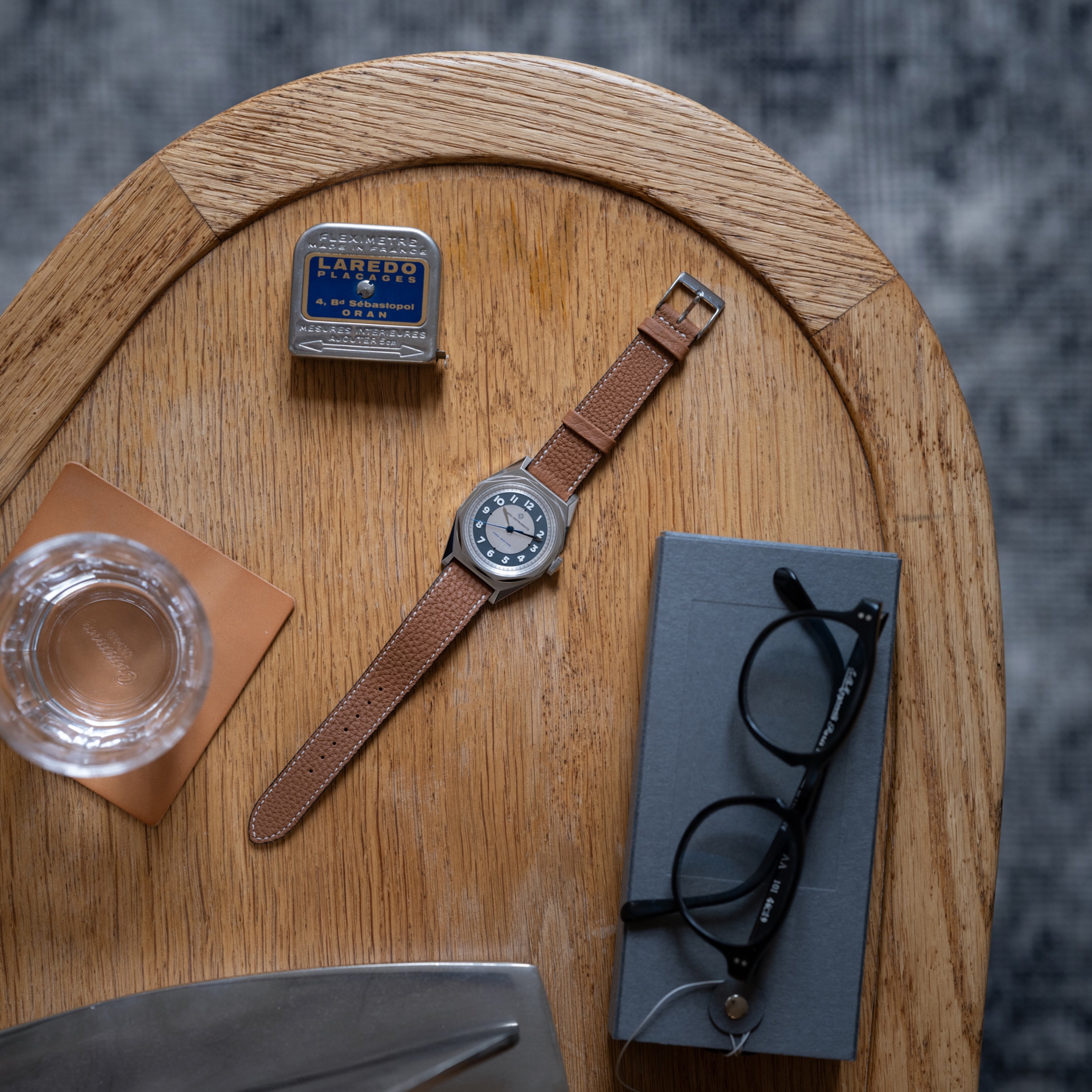 A Merci INSTRUMENTS Beaumarchais H02 Limited Edition For Hodinkee wristwatch, glasses, glass of water, Laredo mints, and a closed notebook on a wooden table.