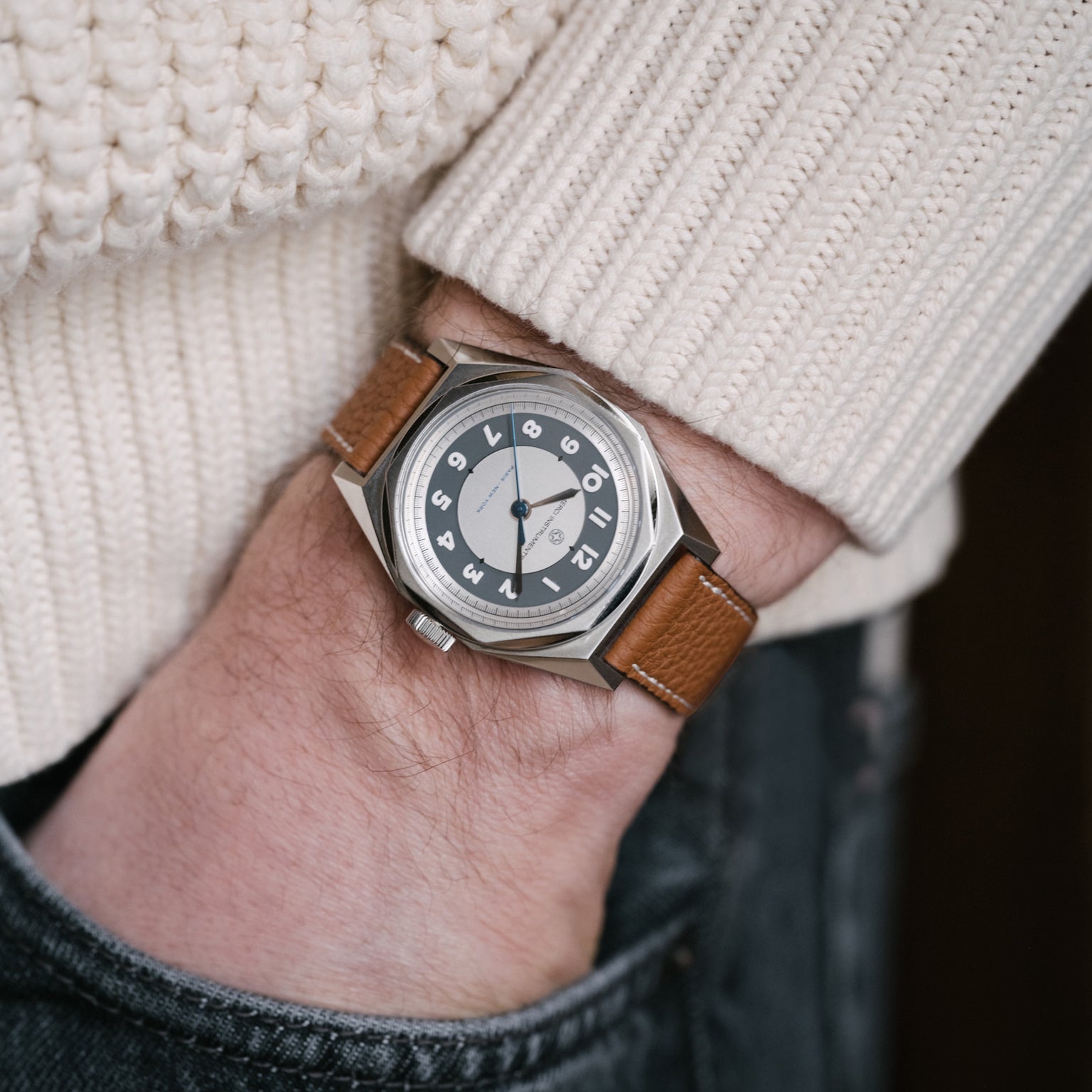 A person wearing the Merci INSTRUMENTS Beaumarchais H02 Limited Edition For Hodinkee by Merci—a silver wristwatch with brown leather strap—hand in denim pocket, dressed in a white sweater.