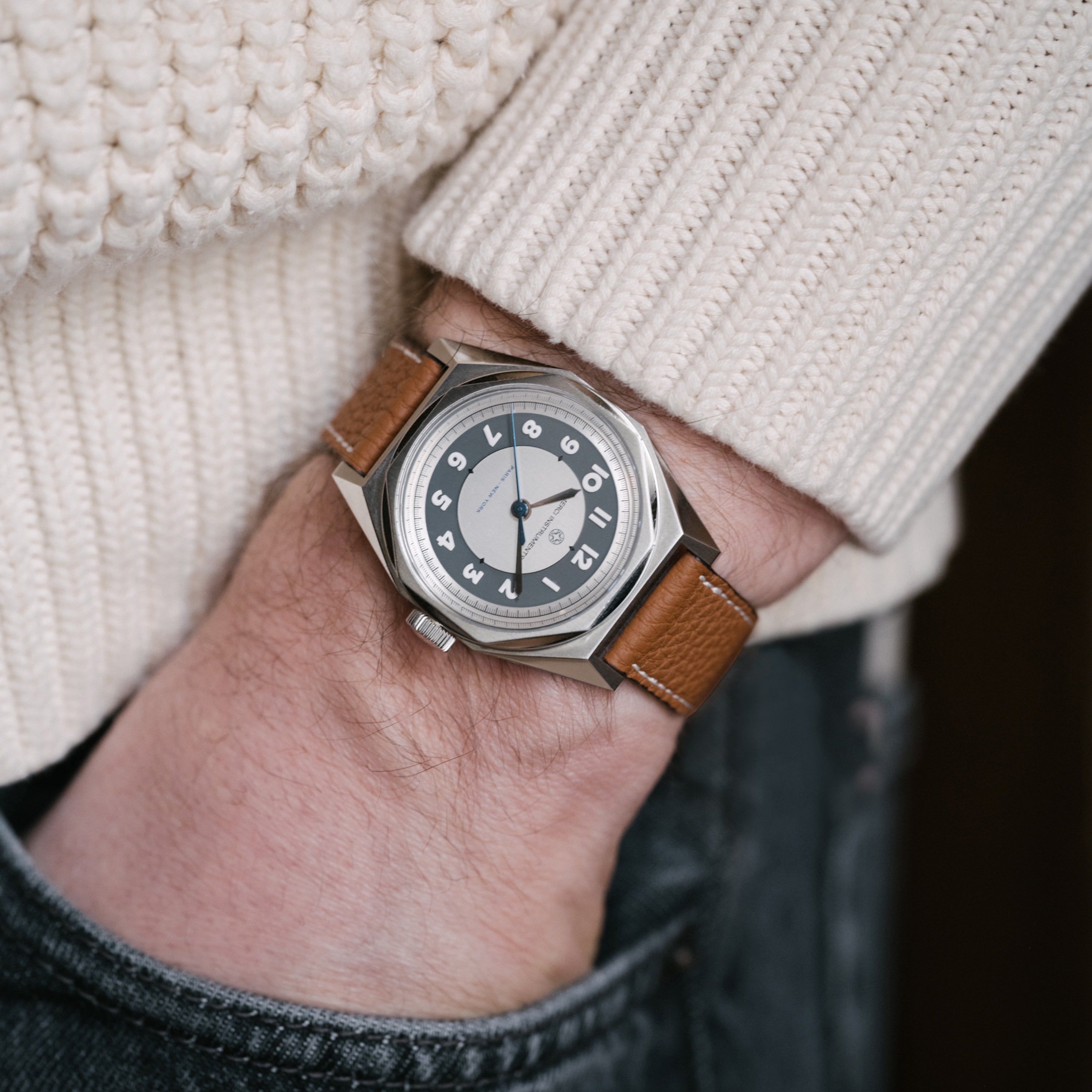 A person wearing the Merci INSTRUMENTS Beaumarchais H02 Limited Edition For Hodinkee by Merci—a silver wristwatch with brown leather strap—hand in denim pocket, dressed in a white sweater.