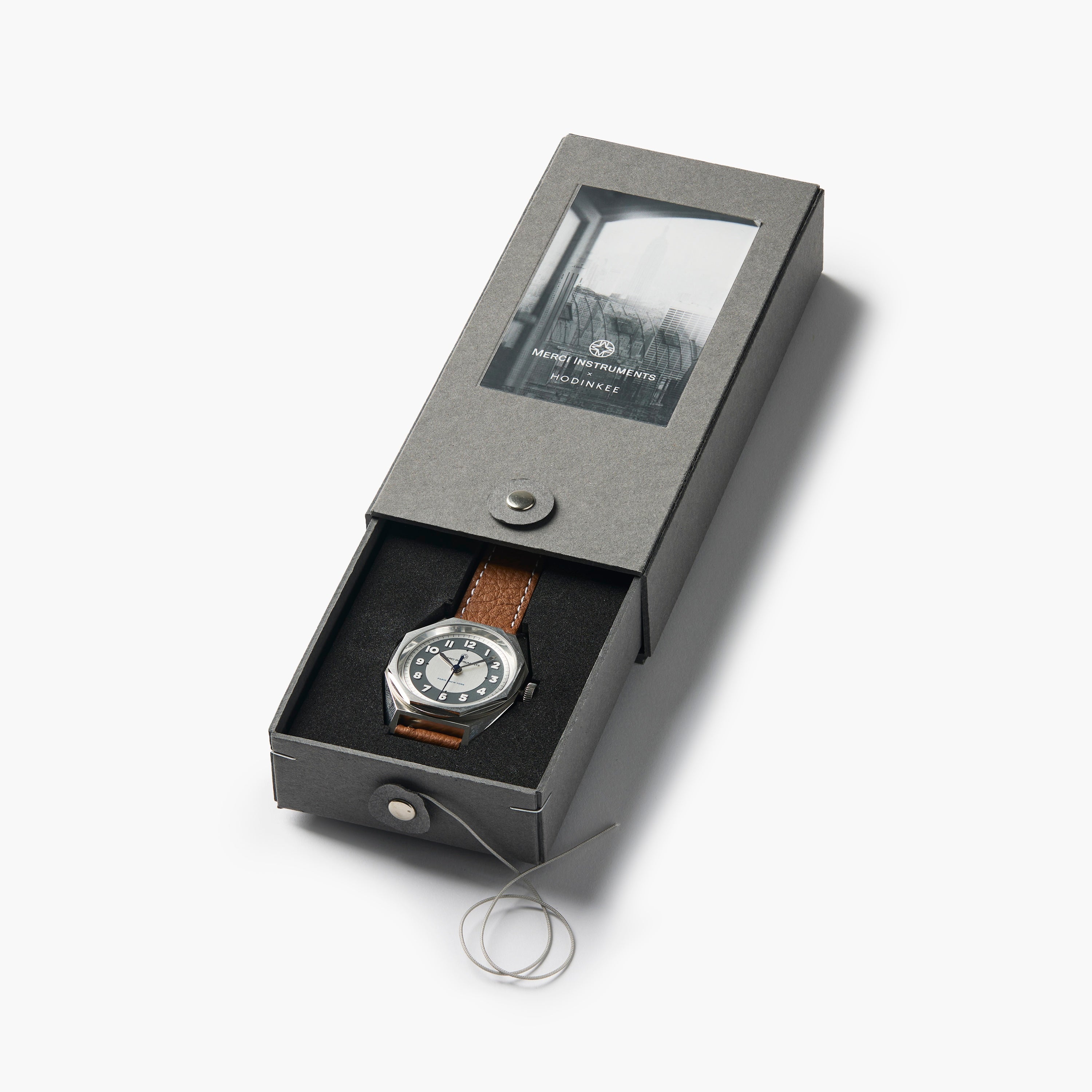 The Merci INSTRUMENTS Beaumarchais H02 Limited Edition For Hodinkee wristwatch by Merci features a brown leather strap and is shown in a gray, partially opened box on a white background.