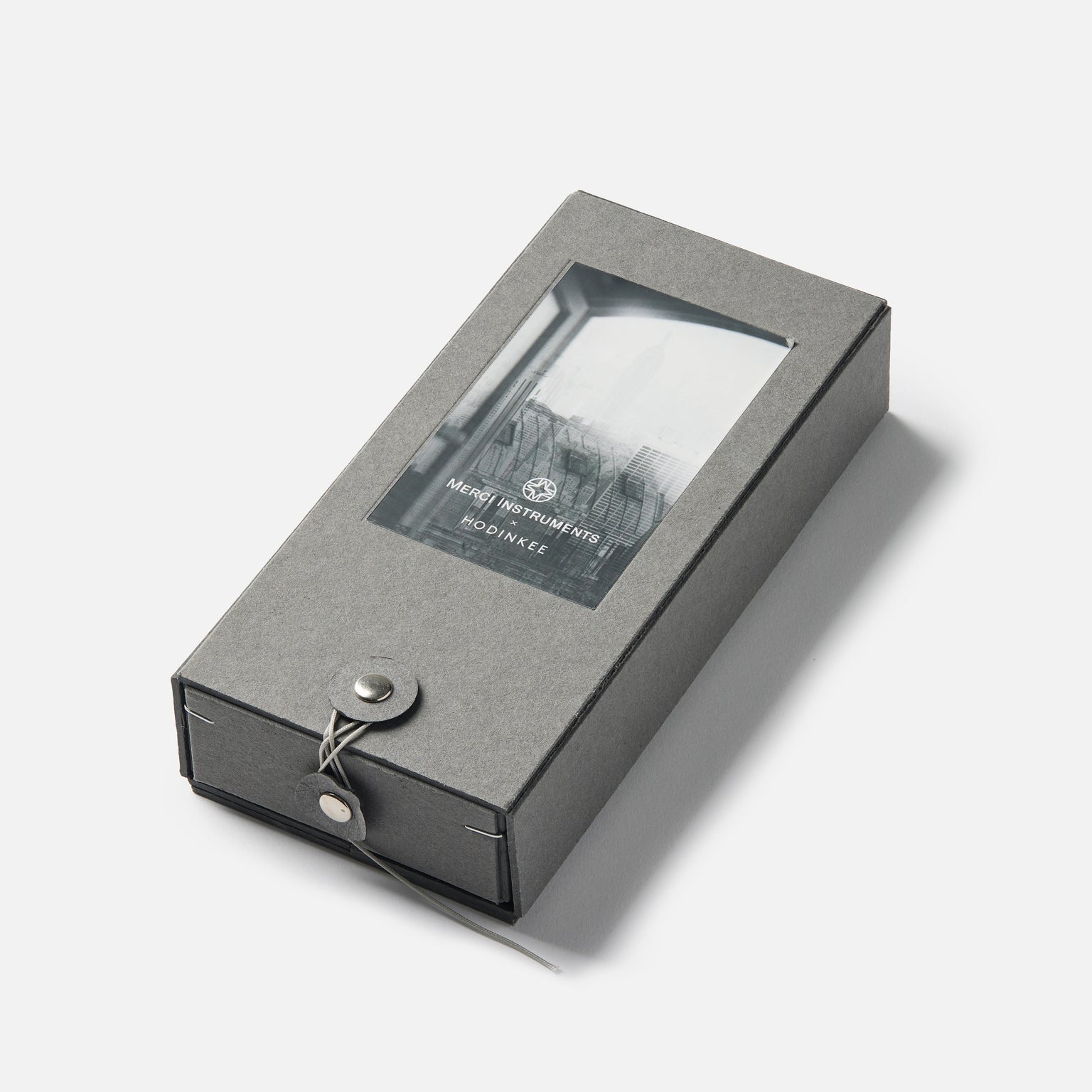 Gray rectangular gift box with string closure and a transparent window on top displaying the Merci INSTRUMENTS Beaumarchais H02 Limited Edition For Hodinkee watch by Merci inside.