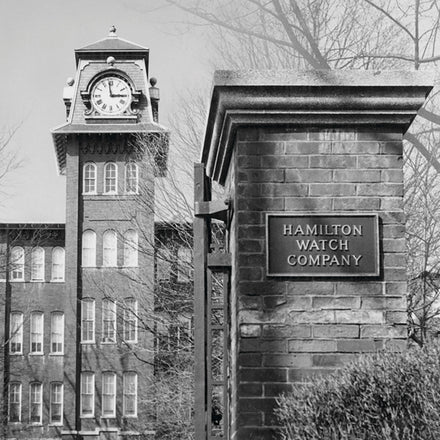 A black and white image of a brick building with a clock tower, a sign on the gatepost reads Hamilton Watch Company