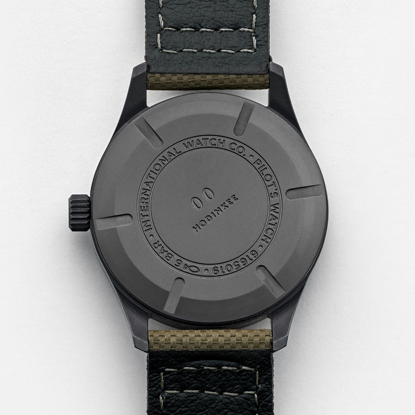 The image shows the back of a black IWC Schaffhausen Pilots Watch Mark XVIII Edition HODINKEE with a leather strap, engraved text around the case, and a central circular logo, highlighting its limited edition status.