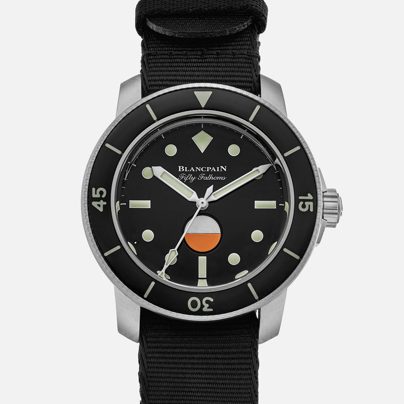 The Blancpain Fifty Fathoms MIL-SPEC Limited Edition for HODINKEE features a black dial, black bezel, luminous markers, orange and white detail above 6 o’clock, and comes on a black fabric strap.