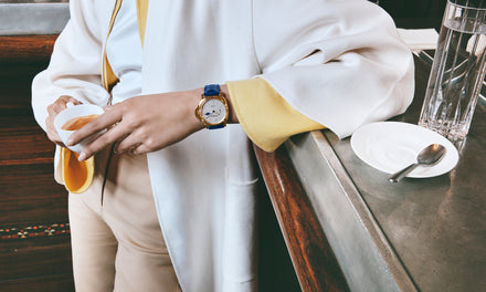 Person in a white and yellow coat holding a cup, wearing a gold watch, standing by a counter with a glass and saucer.