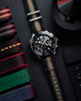 Stainless steel chronograph watch with black dial, white hour markers, and striped fabric strap with red and gold accents.