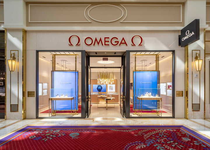 Exterior view of an Omega watch store with glass doors, display windows, and a red carpet at the entrance.