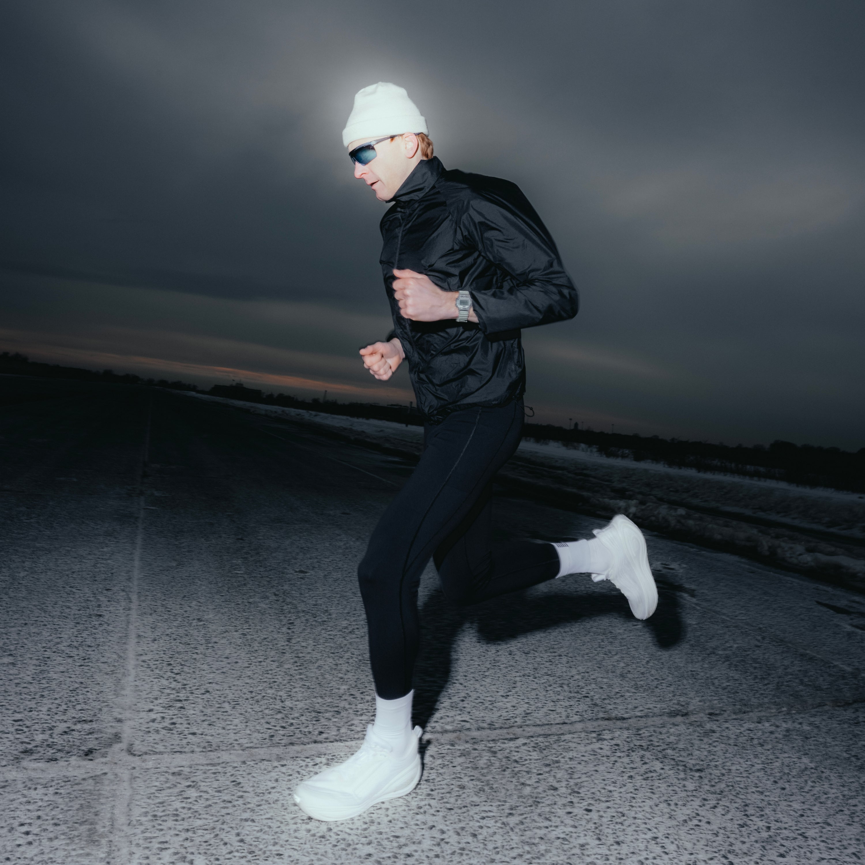 Person jogging at dusk wearing Timex Ironman watch on left wrist, black jacket, white beanie, and sunglasses.