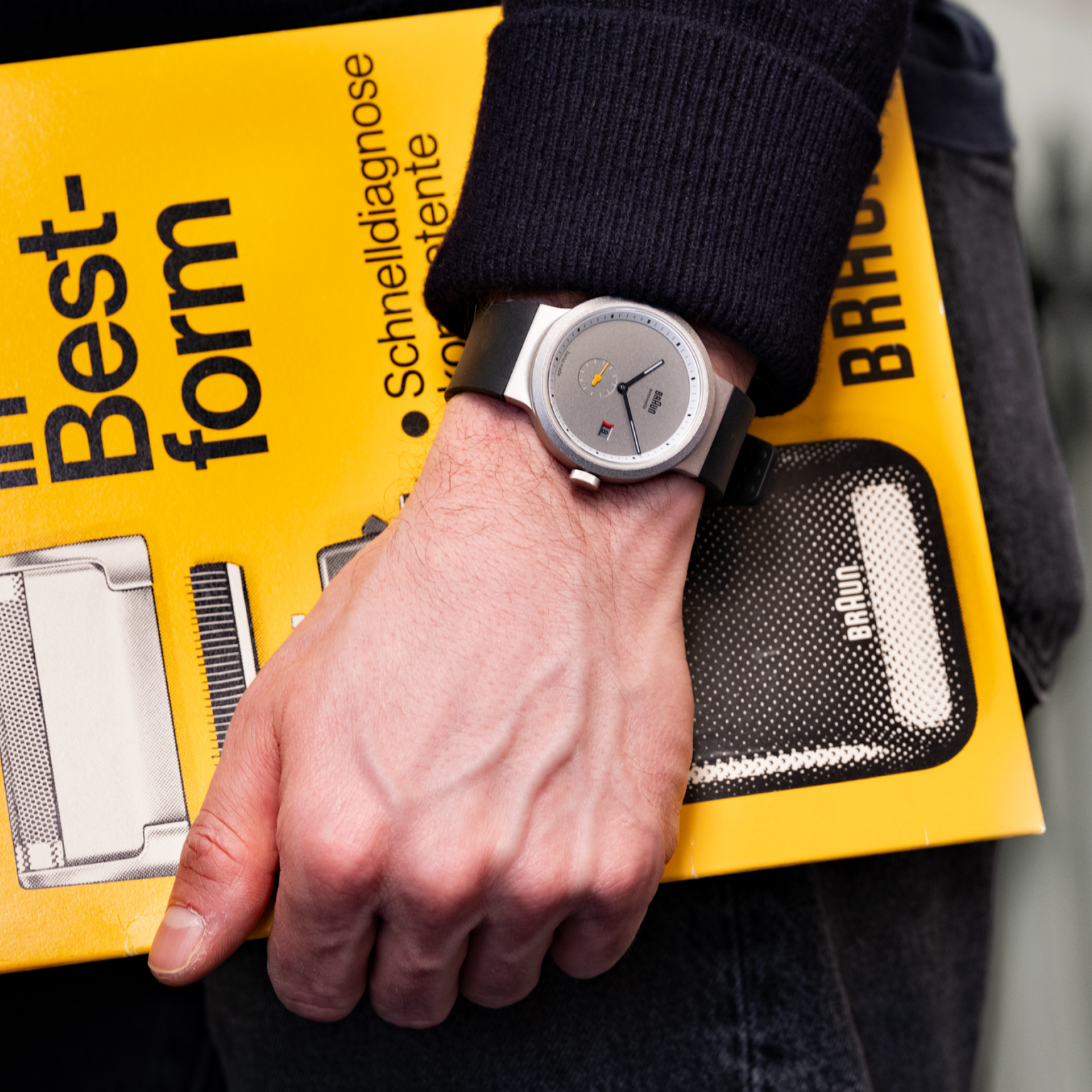 Someone wearing a gray watch holds the Braun BN0279 Sub-Seconds Limited Edition For Hodinkee catalog by their side.