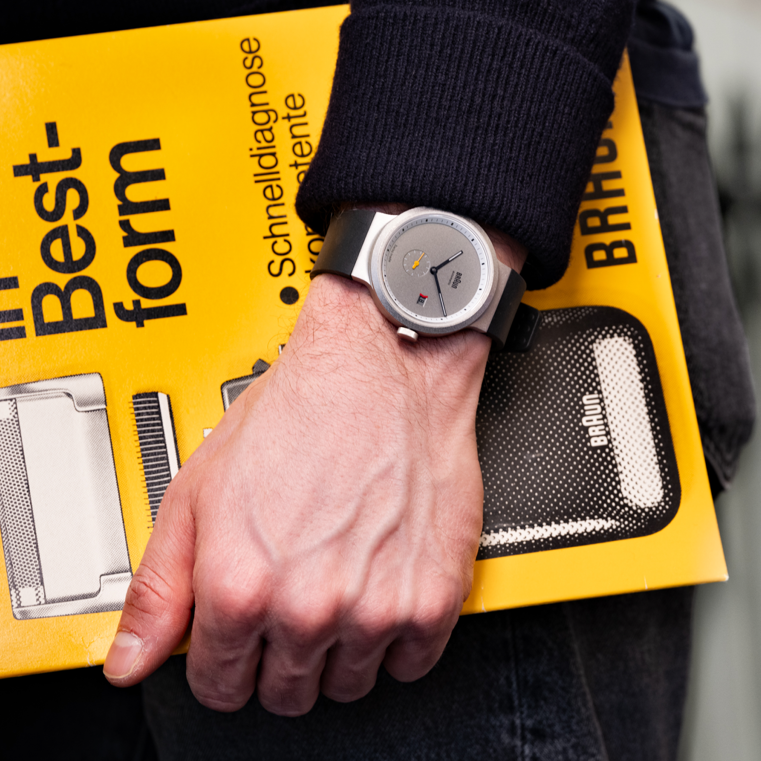 Someone wearing a gray watch holds the Braun BN0279 Sub-Seconds Limited Edition For Hodinkee catalog by their side.