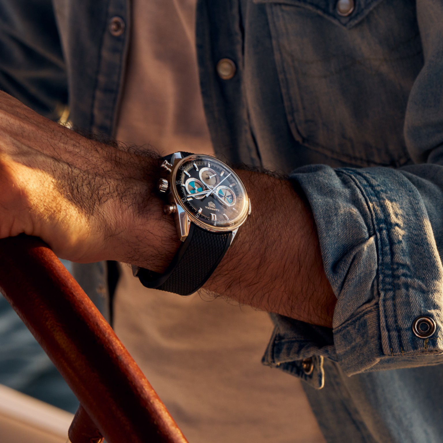 Close-up of a person in a denim shirt wearing the TAG Heuer Carrera Chronograph Seafarer × Hodinkee wristwatch while holding a wooden steering wheel.