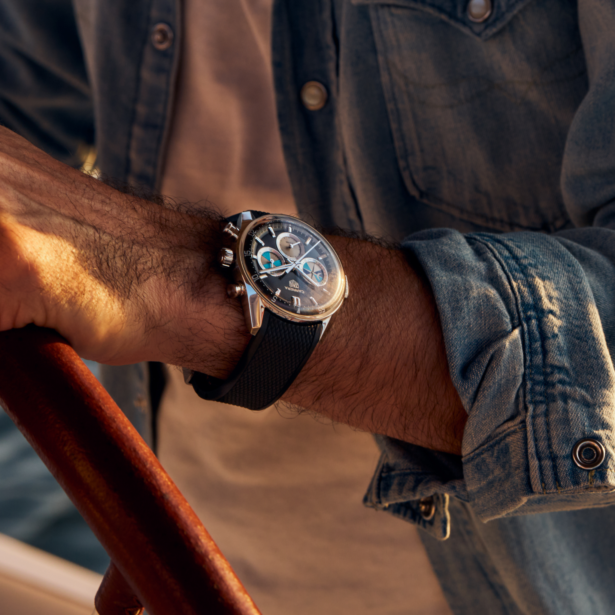 Close-up of a person in a denim shirt wearing the TAG Heuer Carrera Chronograph Seafarer × Hodinkee wristwatch while holding a wooden steering wheel.