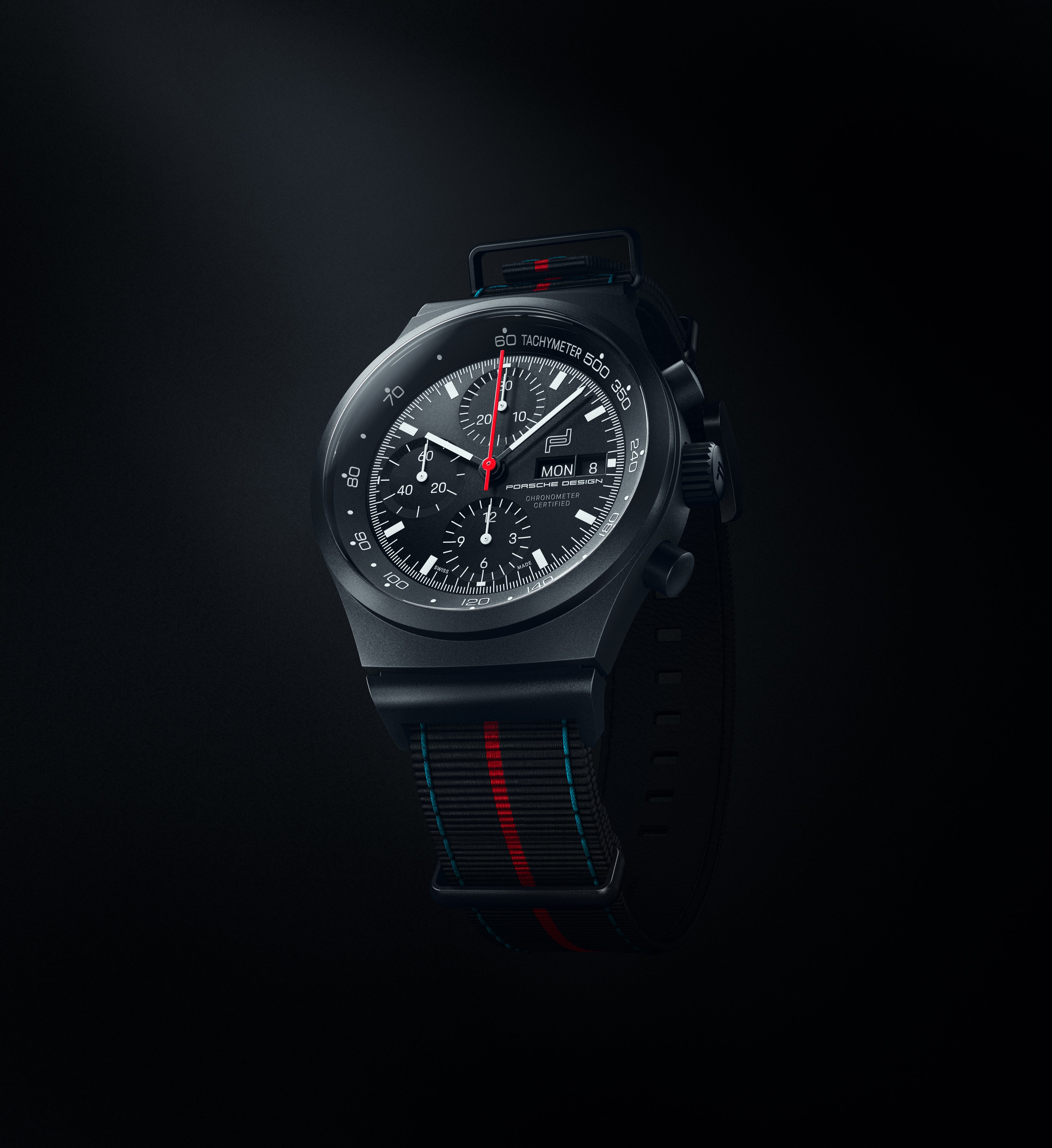 The Chronograph 1 75 Years Porsche Edition by Porsche Design is a black watch with a black strap featuring a red and blue stripe, set against a dark background.