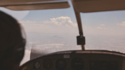 View from inside a small airplane cockpit, looking out at mountains and clouds in the distance.