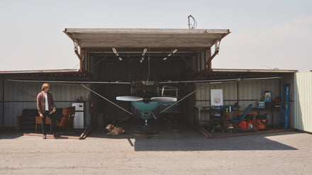 A man and a dog stand outside an open hangar with a small airplane and various equipment inside.