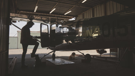 Silhouette of a person entering a small airplane in a hangar, with a dog lying on the floor nearby.