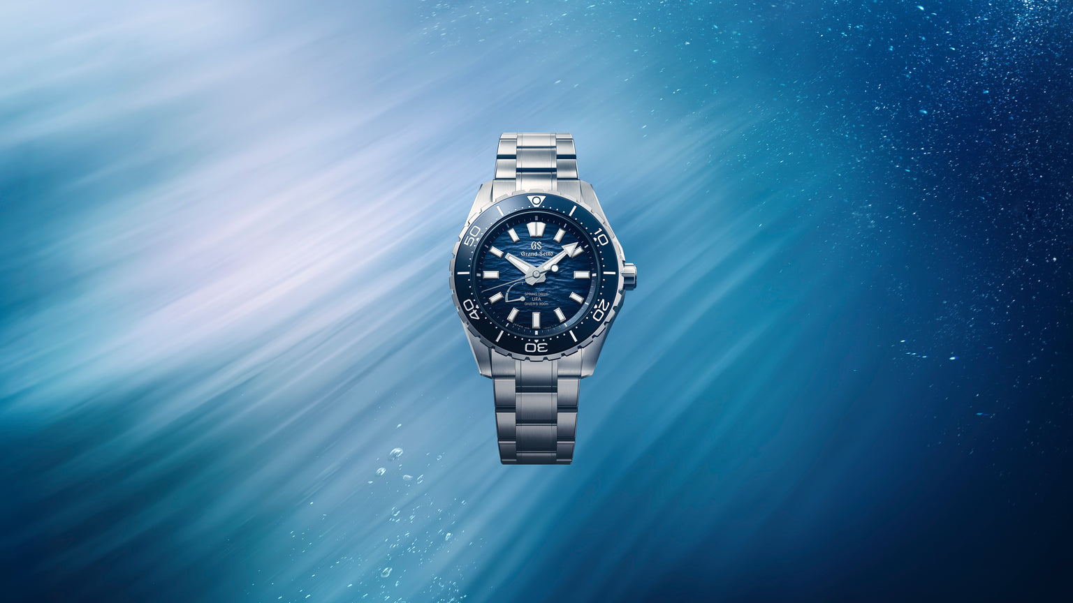 Stainless steel dive watch with dark blue dial, luminous hour markers, and matching metal bracelet on blue water background.