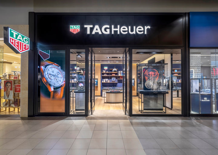 Exterior view of a TAG Heuer store with watches displayed and illuminated brand signage above the entrance.