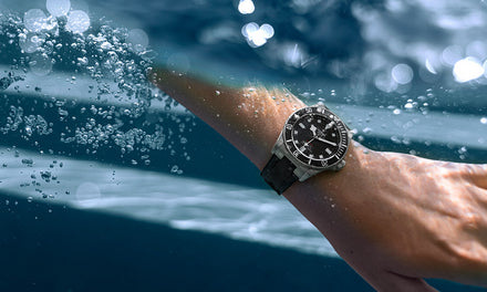 A hand extends into water, viewed from underwater the wrist wears a Tudor Pelagos watch with a black strap and dial