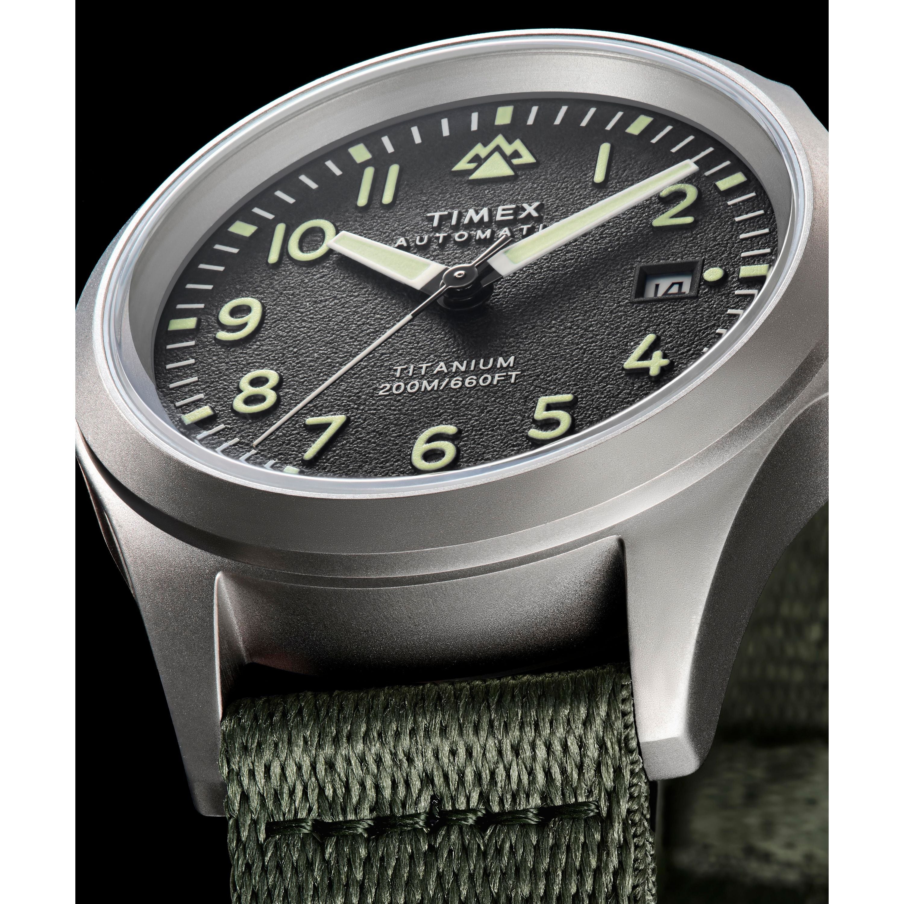 Close-up of the Timex Expedition Titanium Automatic watch with a black textured dial, luminous hands and numerals, date window, titanium 200M/660FT text, Japanese automatic movement, green fabric strap, and silver case.