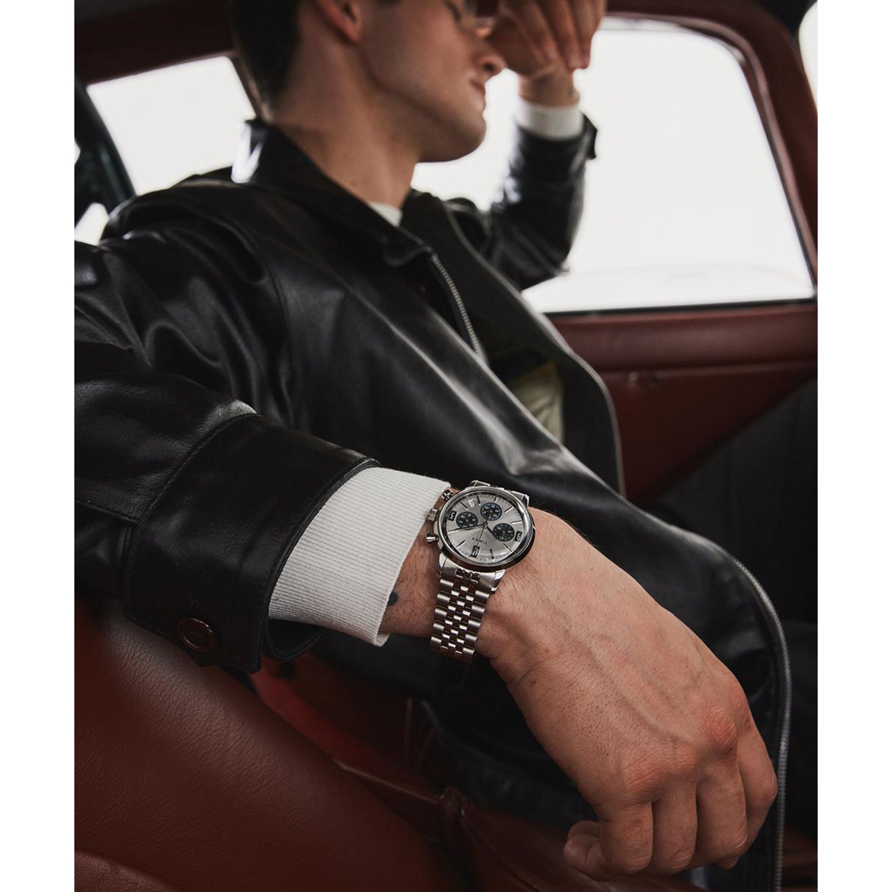 A person in a black leather jacket and sunglasses sits in a car, leaning back with one arm on the door, showcasing a Timex Marlin® Chronograph Tachymeter watch with sleek quartz movement on their wrist.