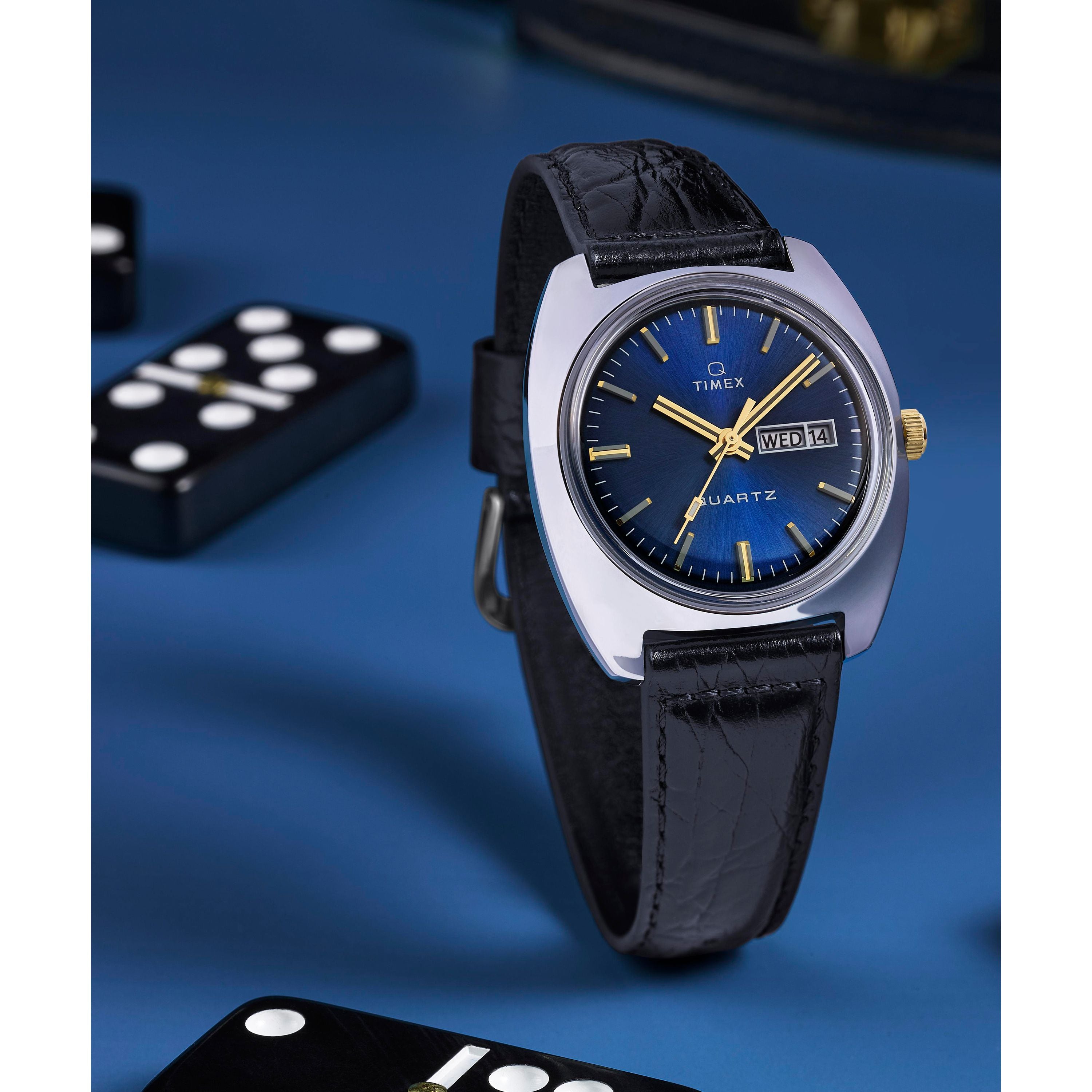 A Timex G SS QZ SW 1978 DAY DTE 37.5 BLU DL 25 watch with a black leather strap and blue sunray dial showing day and date, displayed on a blue surface with domino pieces around it.
